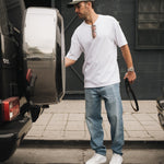 A man stands outdoors near the back of a black SUV wearing a white t-shirt, a cap, and The Jeans Relaxed Straight Fit in Light Midwash by c'est normal.