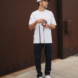 A man stands outdoors holding a camera, wearing The Jeans Relaxed Straight Fit in rinse wash, paired with a white t-shirt, white sneakers, and a cap by c'est normal.