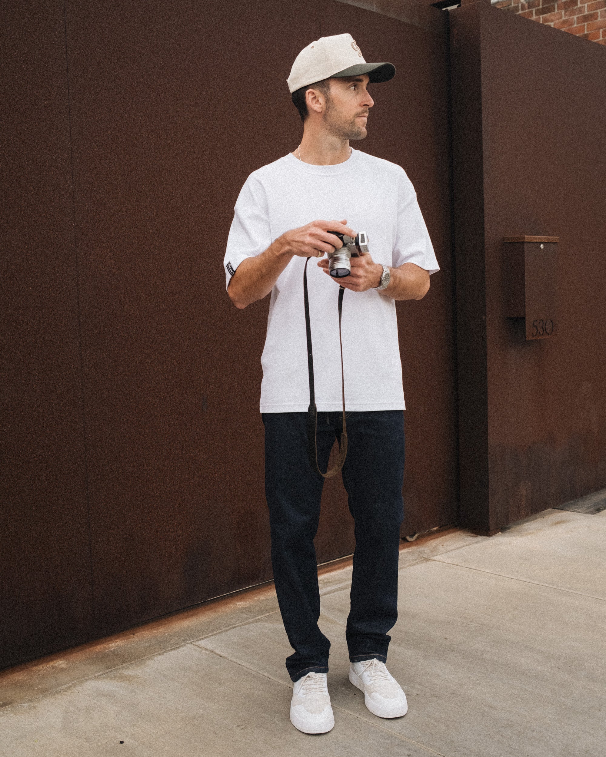 A man stands outdoors holding a camera, wearing The Jeans Relaxed Straight Fit in rinse wash, paired with a white t-shirt, white sneakers, and a cap by c'est normal.