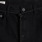 A close-up shot of the front waist of The Jeans Slim Fit in Jet Black, highlighting the branded button, front pockets, subtle orange accent stitching, and a knotted black cord. A white c'est normal label is visible inside the waistband.