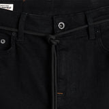 A close-up shot of the front waist of The Jeans Slim Fit in Jet Black, highlighting the branded button, front pockets, subtle orange accent stitching, and a knotted black cord. A white c'est normal label is visible inside the waistband.