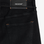 A close-up shot of the back of The Jeans Slim Fit in Jet Black, laid flat on a white background. The image highlights the back pocket with subtle orange accent stitching and the black rubber patch at the back waist featuring the white c'est normal logo.