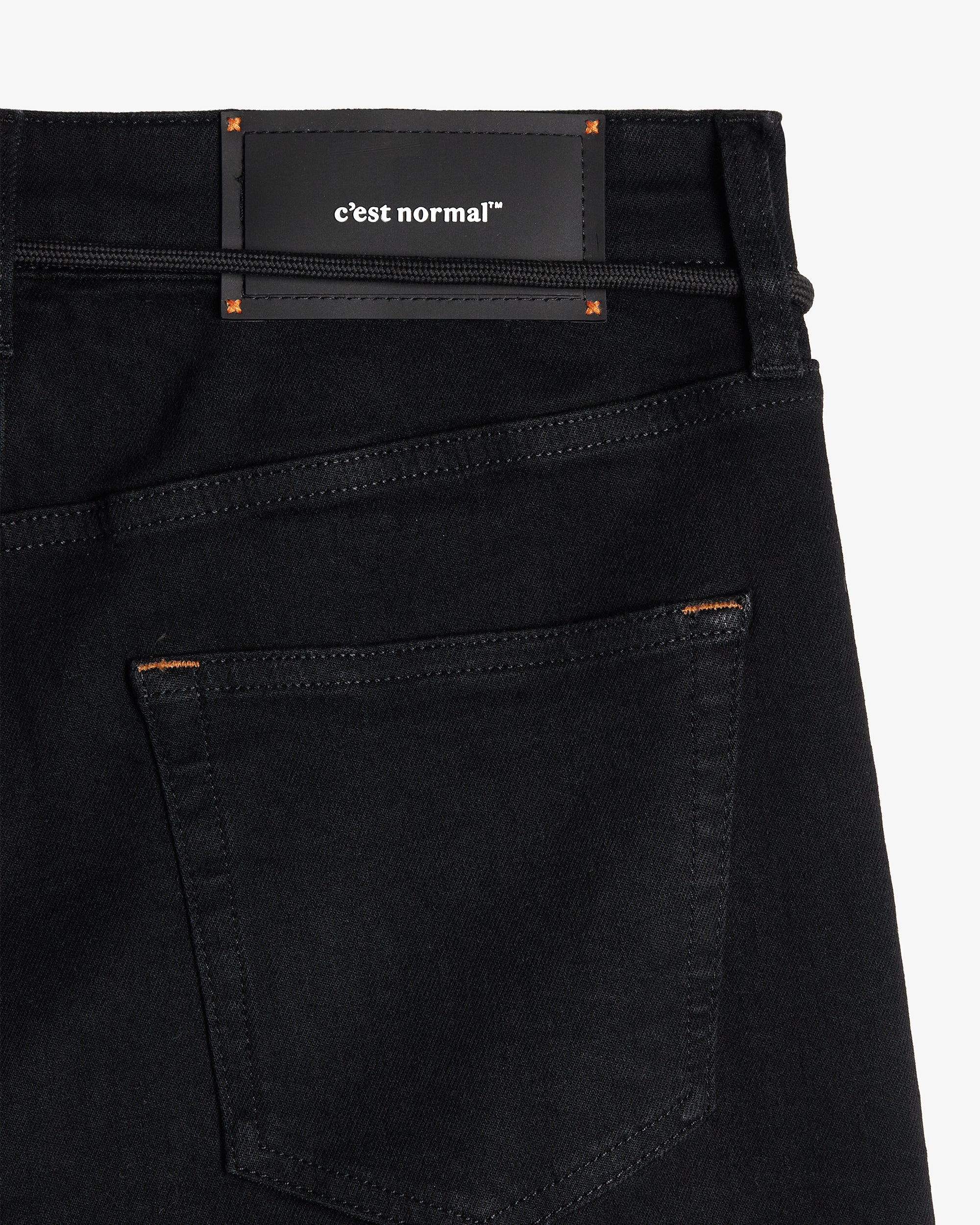 A close-up shot of the back of The Jeans Slim Fit in Jet Black, laid flat on a white background. The image highlights the back pocket with subtle orange accent stitching and the black rubber patch at the back waist featuring the white c'est normal logo.