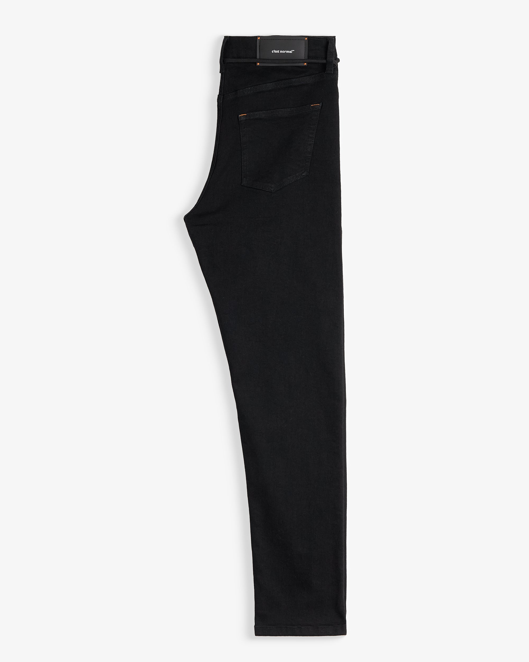 The Jeans Slim Fit in Jet Black, folded in half and laid flat on a white background. The image highlights the back pocket, subtle orange accent stitching, and the rubber patch with the c'est normal logo at the back waist.