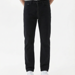 A model is wearing The Jeans Slim Fit by c'est normal in Jet Black, paired with a white t-shirt and white sneakers, standing against a white background. The image highlights the clean slim silhouette and classic denim look.