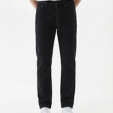 A model is wearing The Jeans Slim Fit by c'est normal in Jet Black, paired with a white t-shirt and white sneakers, standing against a white background. The image highlights the clean slim silhouette and classic denim look.