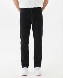 A model is wearing The Jeans Slim Fit by c'est normal in Jet Black, paired with a white t-shirt and white sneakers, standing against a white background. The image highlights the clean slim silhouette and classic denim look.