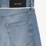 A close-up view of the back of The Jeans Slim Fit in Light Midwash, highlighting the back pocket, the black rubber patch at the back waist with the c'est normal logo, and the subtle orange accent stitching.