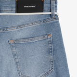 A close-up view of the back of The Jeans Slim Fit in Light Midwash, highlighting the back pocket, the black rubber patch at the back waist with the c'est normal logo, and the subtle orange accent stitching.
