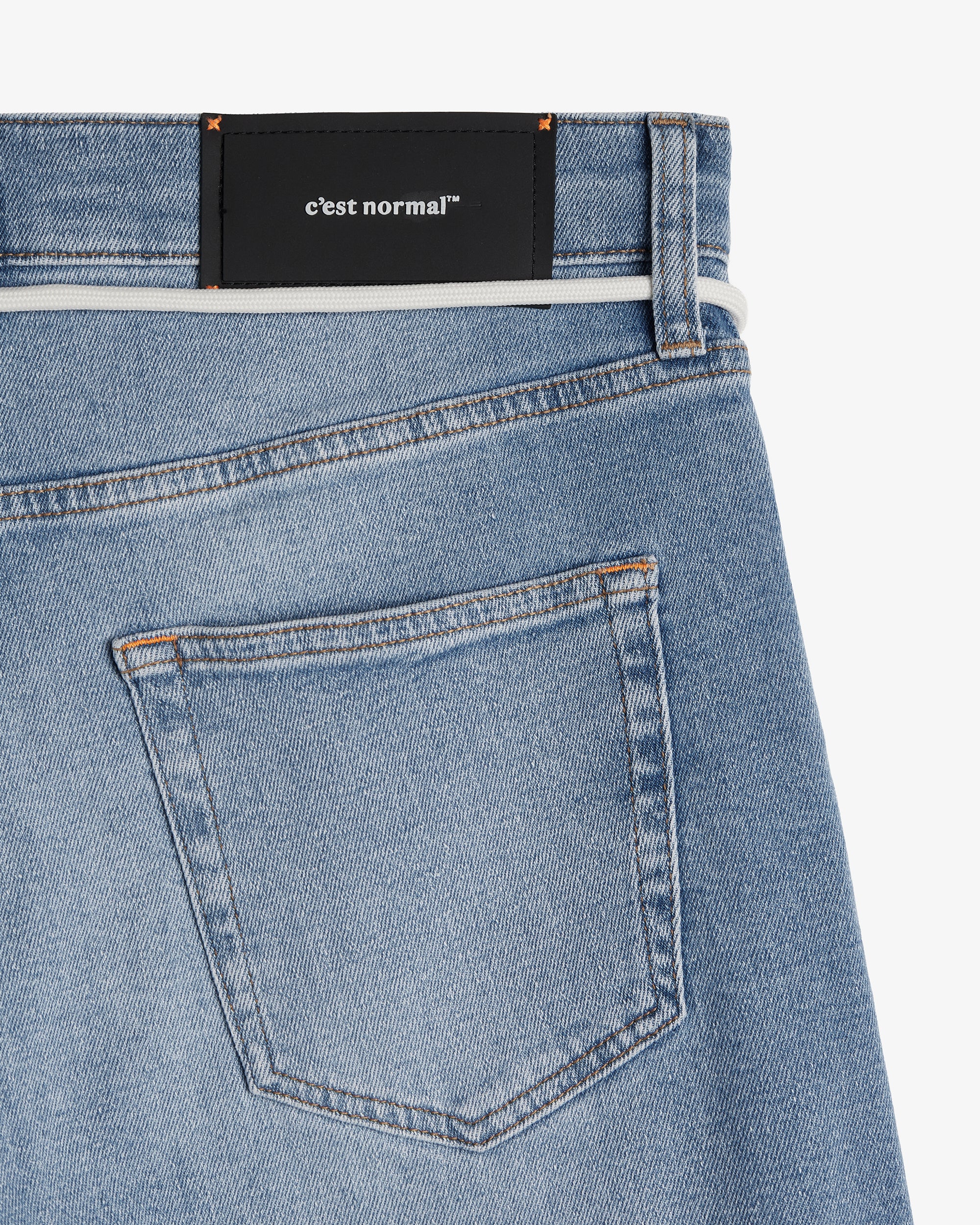 A close-up view of the back of The Jeans Slim Fit in Light Midwash, highlighting the back pocket, the black rubber patch at the back waist with the c'est normal logo, and the subtle orange accent stitching.