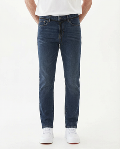 A model standing on a white background wearing The Jeans Slim Fit in a midwash blue color by c'est normal. The image highlights the clean slim leg silhouette, subtle orange accent stitching, and a black drawstring detail at the waist.
