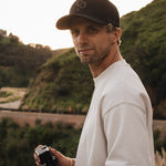 A man wearing The Snapback Cap in black and a white t-shirt stands outdoors holding a camera against a backdrop of green hills. The image highlights the embroidered mirror logo on the front and the c'est normal logo on the side of the cap.
