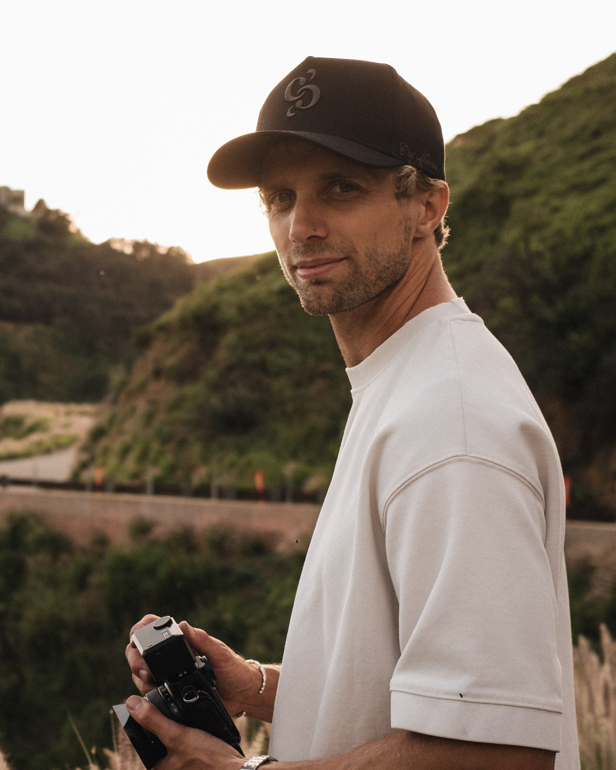 A man wearing The Snapback Cap in black and a white t-shirt stands outdoors holding a camera against a backdrop of green hills. The image highlights the embroidered mirror logo on the front and the c'est normal logo on the side of the cap.