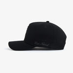 A side view of The Snapback Cap in black, placed on a white background. The image highlights the curved brim and the embroidered c'est normal text on the side panel.