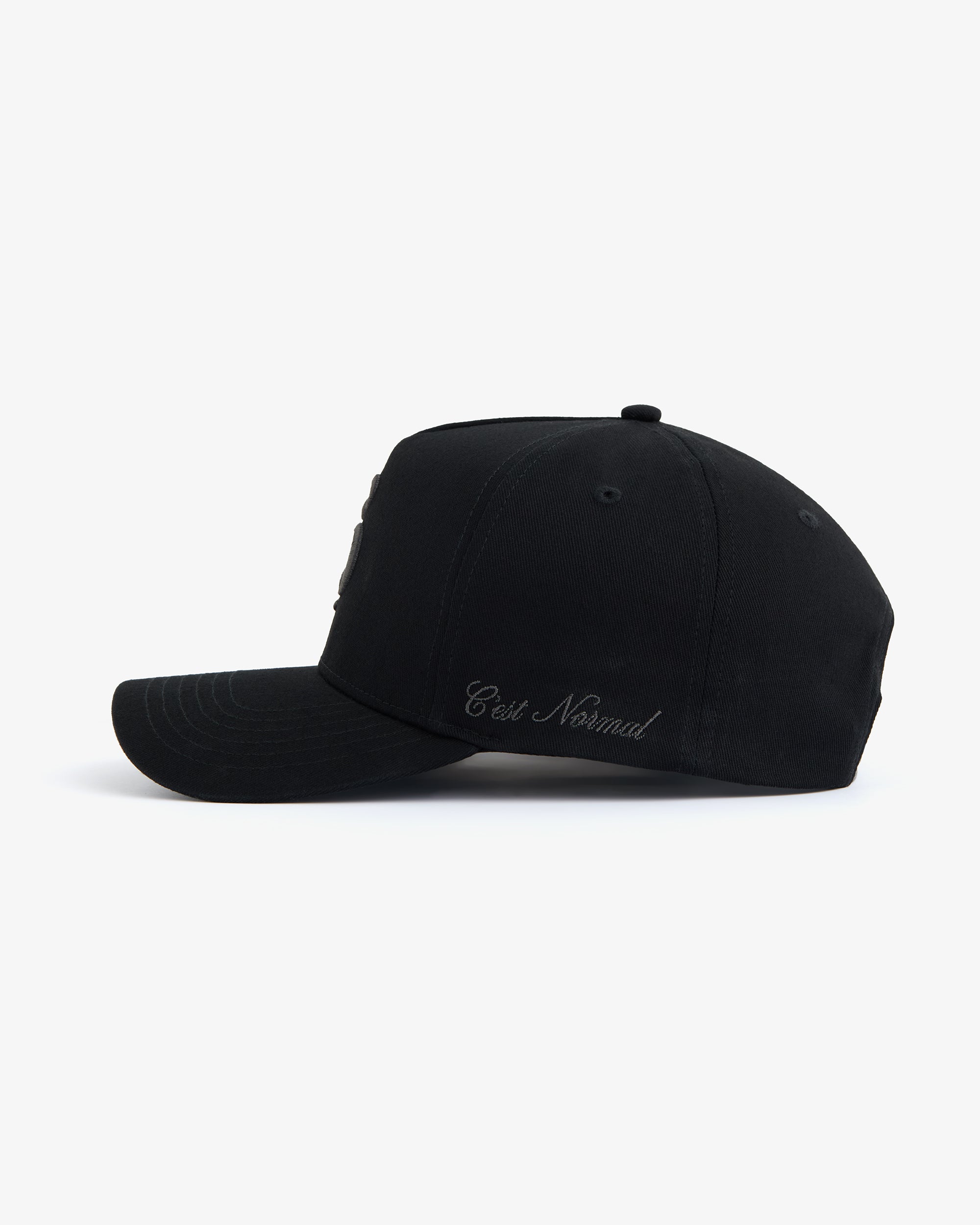 A side view of The Snapback Cap in black, placed on a white background. The image highlights the curved brim and the embroidered c'est normal text on the side panel.