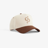 A front view of The Snapback Cap in cream and brown, set against a white background. The image highlights the cream crown, brown brim, and the brown embroidered c'est normal mirror logo on the front.