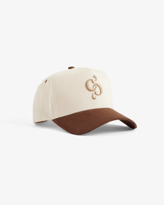 A front view of The Snapback Cap in cream and brown, set against a white background. The image highlights the cream crown, brown brim, and the brown embroidered c'est normal mirror logo on the front.