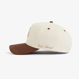 A side view of The Snapback Cap in cream and brown, laid flat on a white background. The image highlights the brown brim, matching eyelets, and the embroidered c'est normal logo on the side.