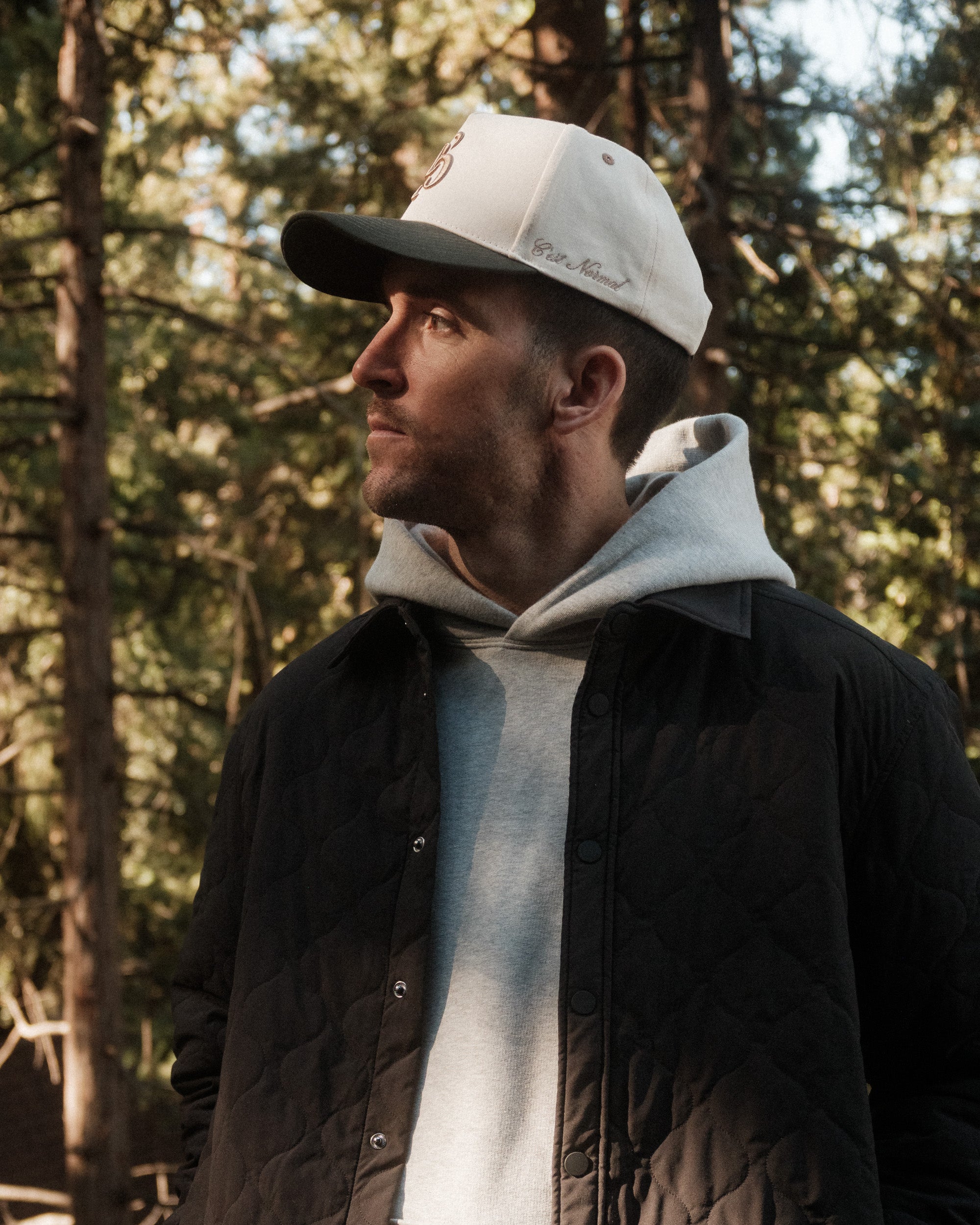 A man looking to the side in a forest setting, wearing The Snapback Cap in Cream/Pine, a grey hoodie, and a black quilted jacket. The cap features an embroidered c'est normal logo on the side.
