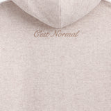 The Toulouse Hoodie in beige — close-up of embroidered C'est normal logo below the hood