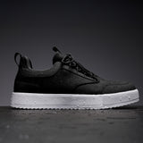 A side profile of The Dynamic Waterproof Sneaker 2.0 Low in black with a white sole, resting on a wet, dark surface. The image highlights the water droplets on the shoe's upper and sole, showcasing its waterproof design by c'est normal.