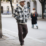 A man wearing The 5 Pocket Pants in brown, paired with a white hoodie, a black and white check jacket, and a c'est normal cap, walking down a city street.