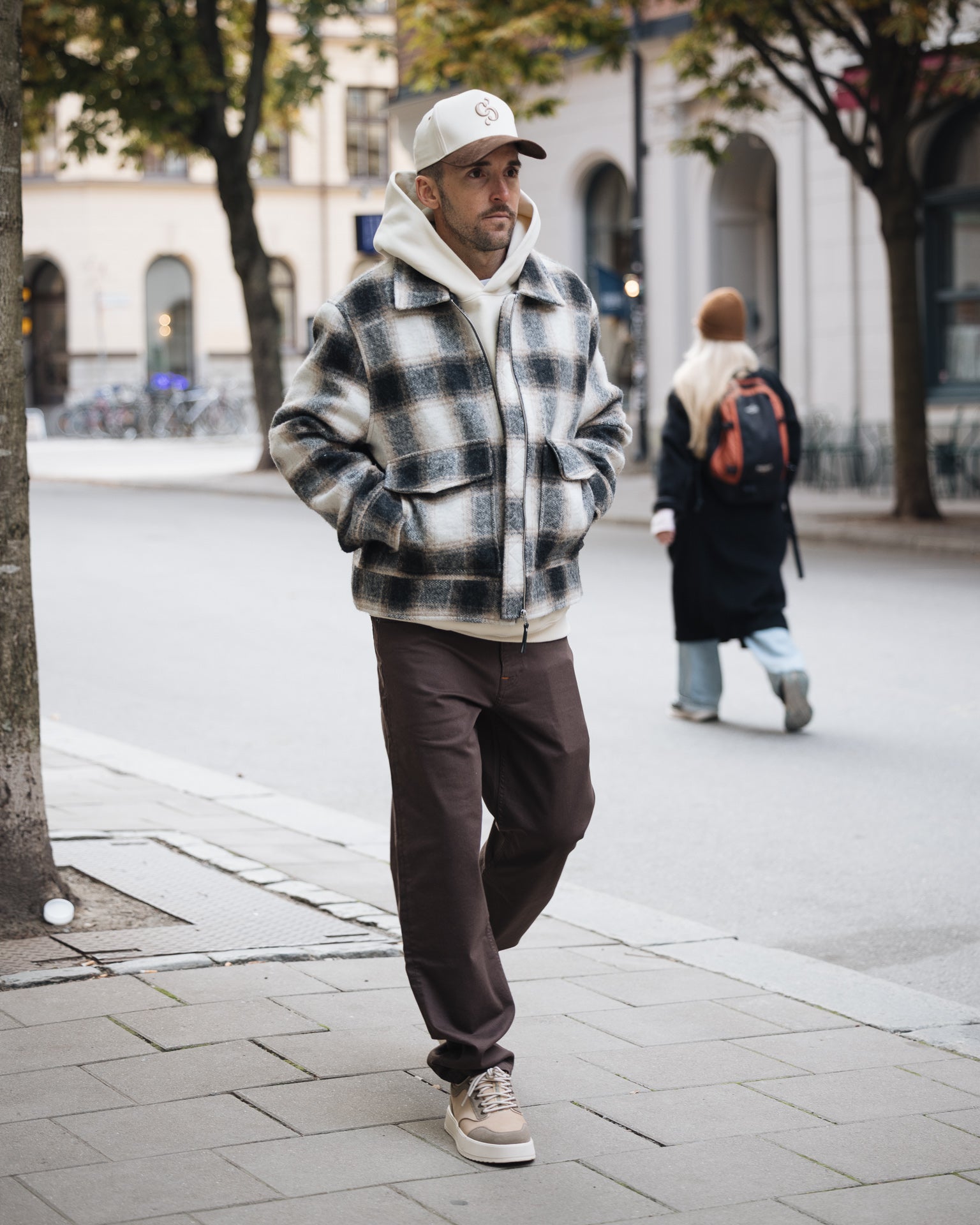 A man wearing The 5 Pocket Pants in brown, paired with a white hoodie, a black and white check jacket, and a c'est normal cap, walking down a city street.