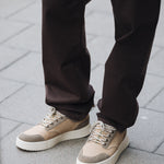 A close-up shot of the lower half of a person wearing The 5 Pocket Pants in brown, standing on a paved sidewalk. The person is also wearing light-colored sneakers.