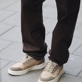 A close-up shot of the lower half of a person wearing The 5 Pocket Pants in brown, standing on a paved sidewalk. The person is also wearing light-colored sneakers.