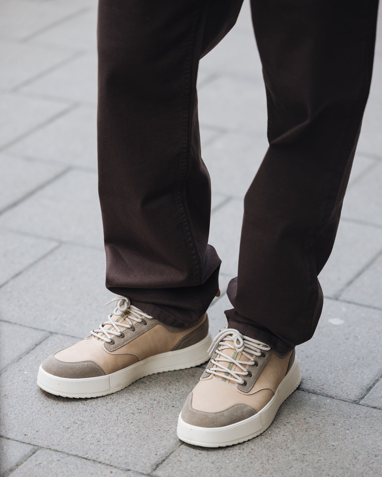 A close-up shot of the lower half of a person wearing The 5 Pocket Pants in brown, standing on a paved sidewalk. The person is also wearing light-colored sneakers.