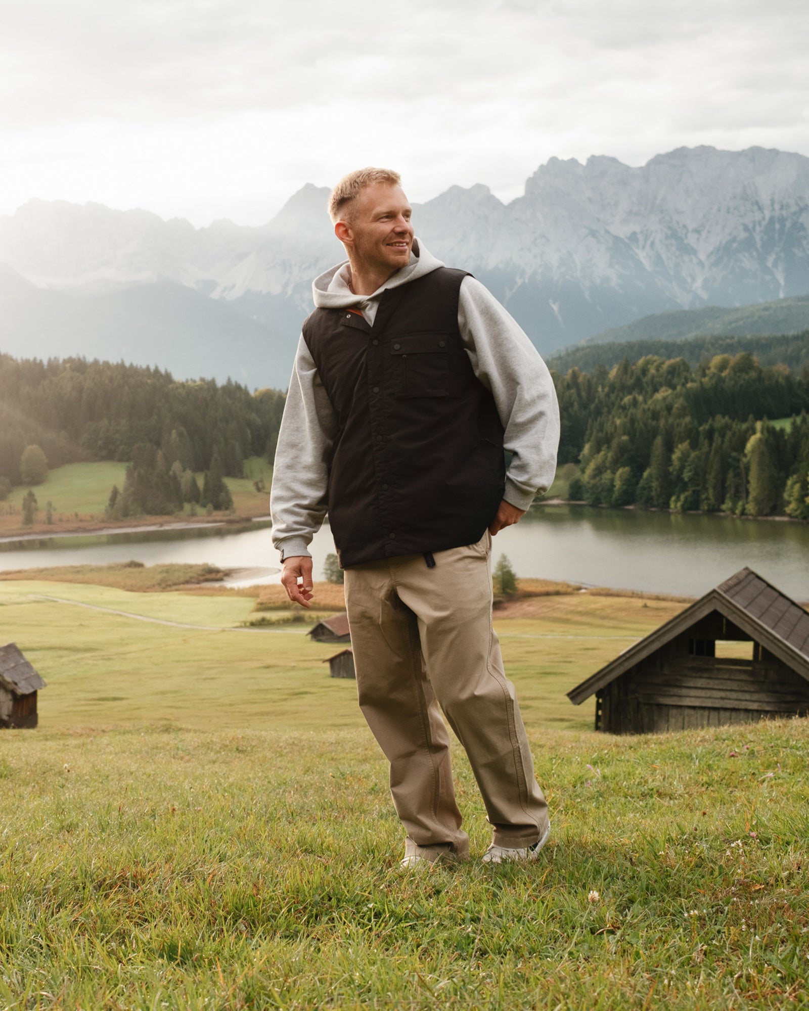 The Utility Vest in black — worn outdoors with a grey hoodie and a mountain backdrop