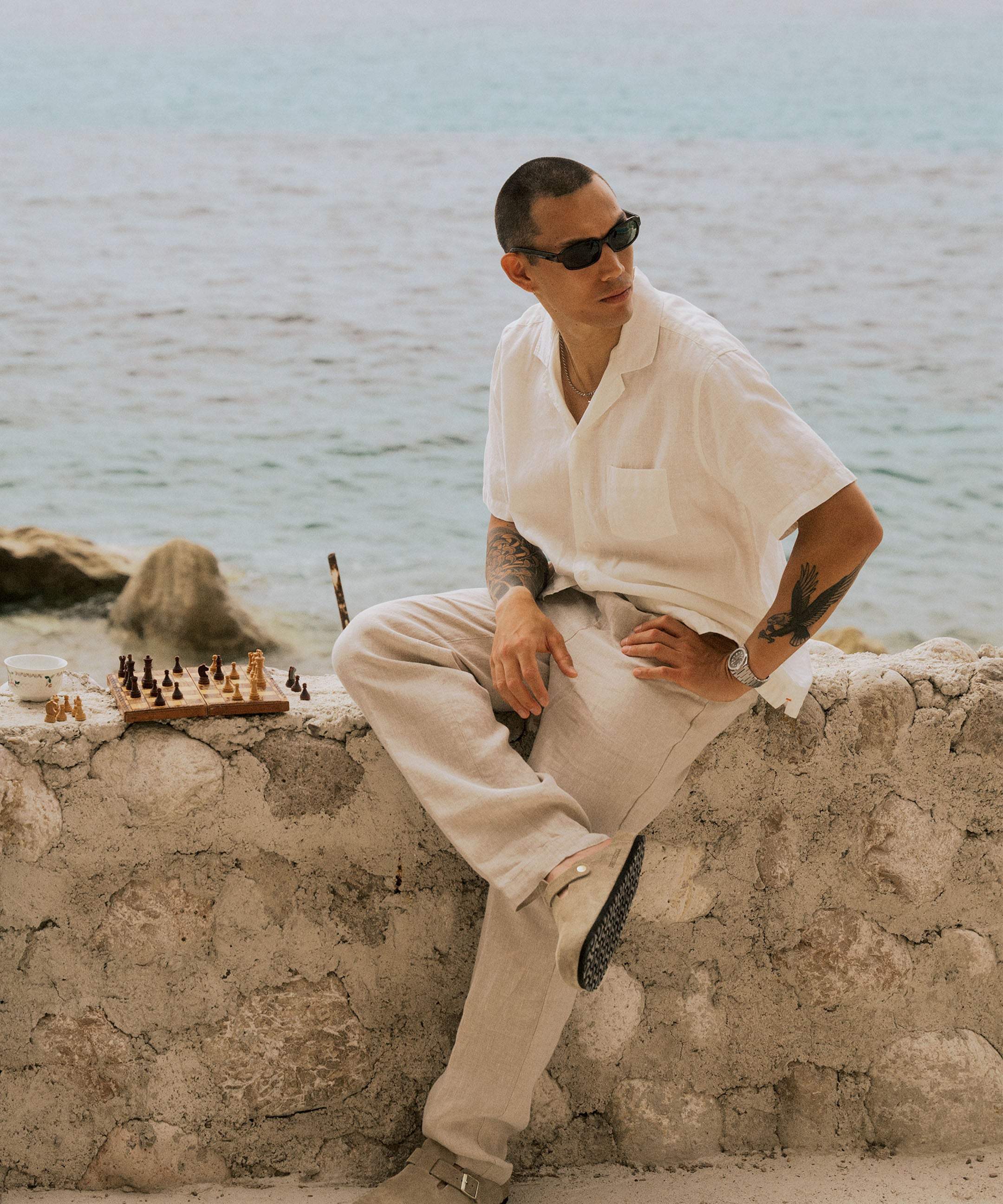 The Holiday Linen Trouser in Natural — sitting on a stone wall next to a chessboard