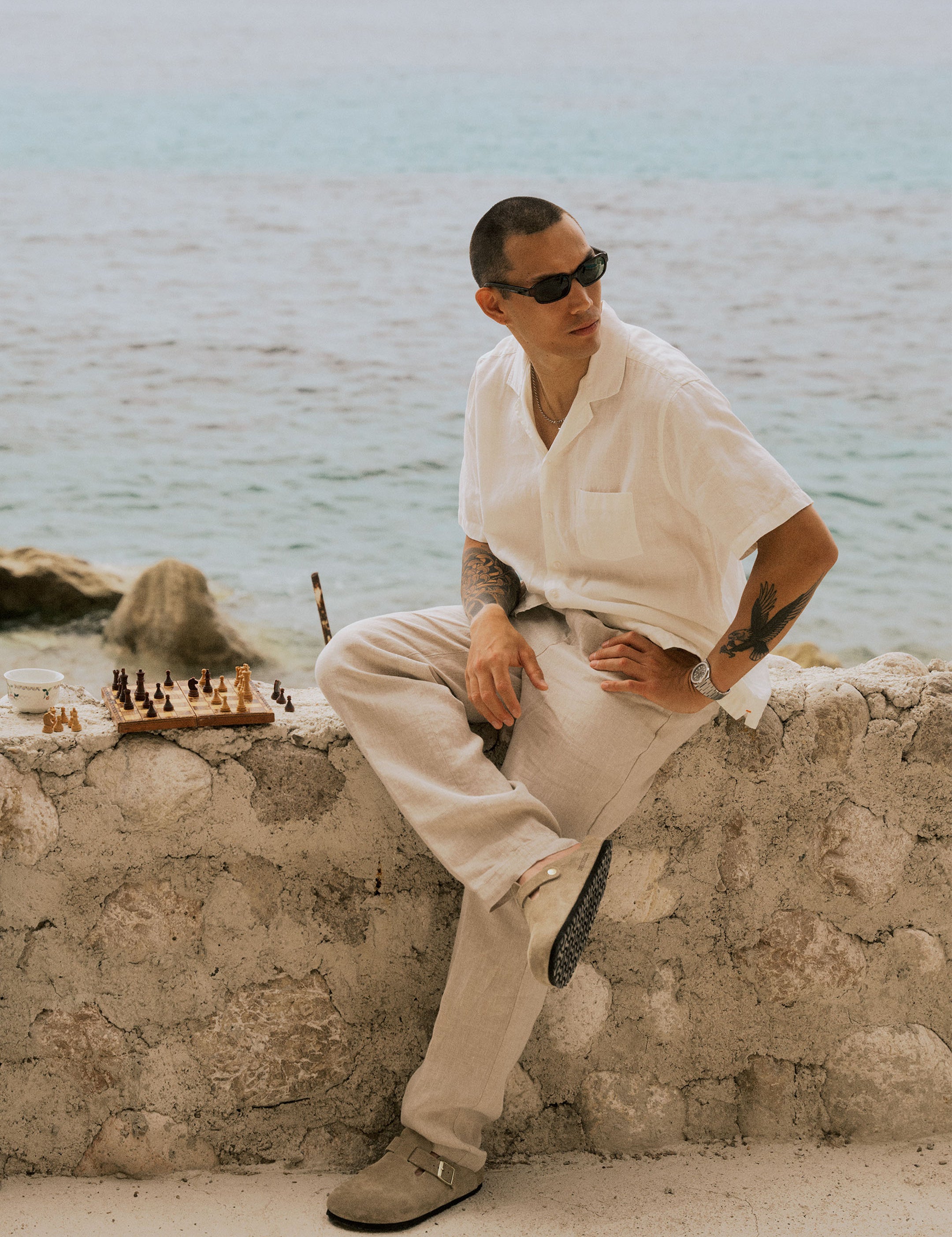 The Holiday Linen Trouser in Natural — sitting on a stone wall next to a chessboard