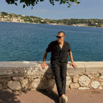 The Holiday Linen Trouser in black — worn leaning against a stone wall by the sea