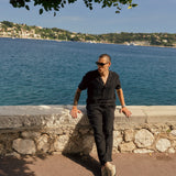 The Holiday Linen Trouser in black — worn leaning against a stone wall by the sea