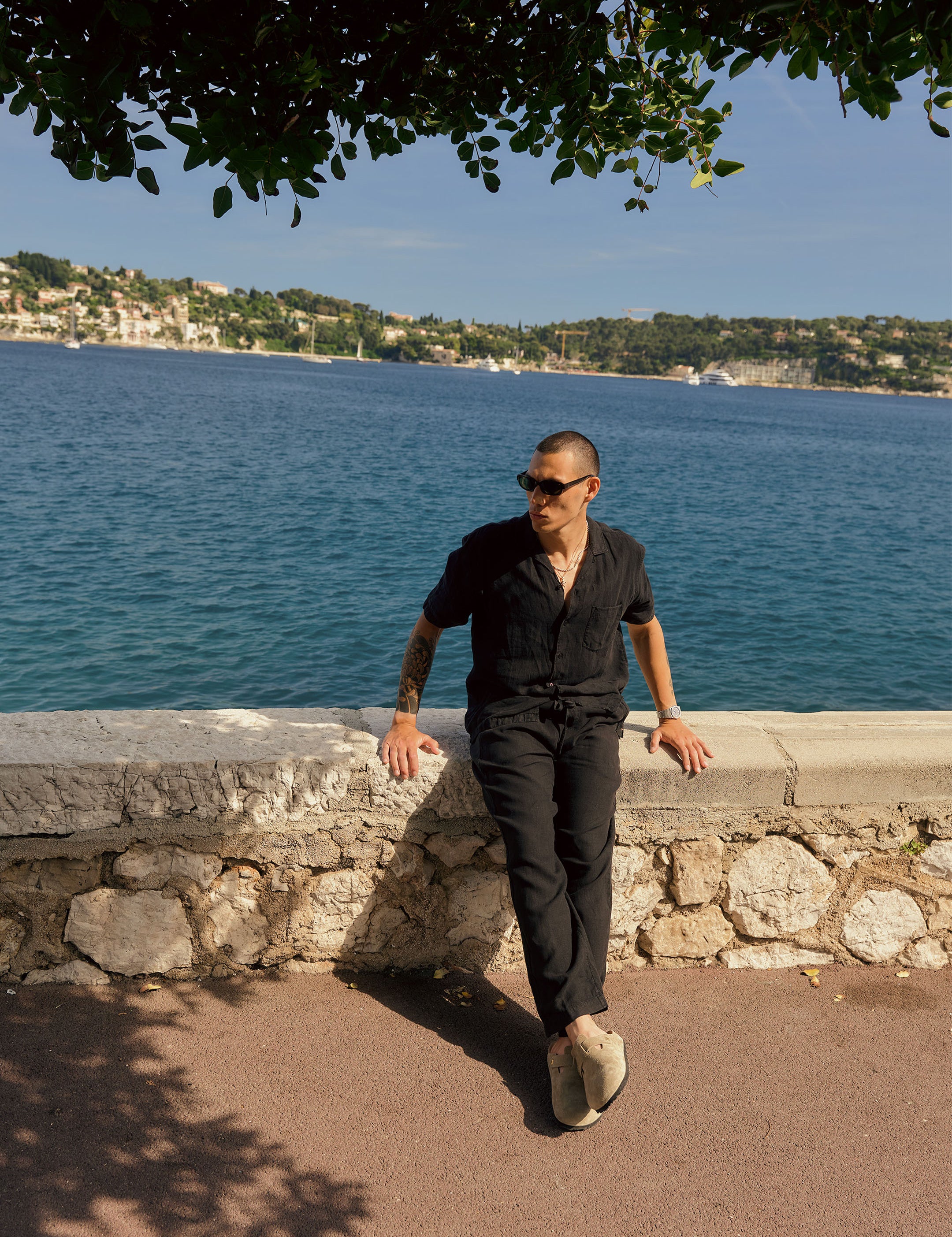 The Holiday Linen Trouser in black — worn leaning against a stone wall by the sea