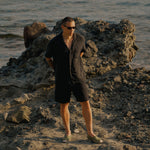 The Holiday Linen SS Shirt in black — worn on a rocky beach by the ocean
