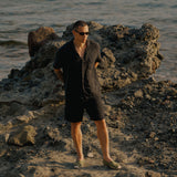 The Holiday Linen SS Shirt in black — worn on a rocky beach by the ocean