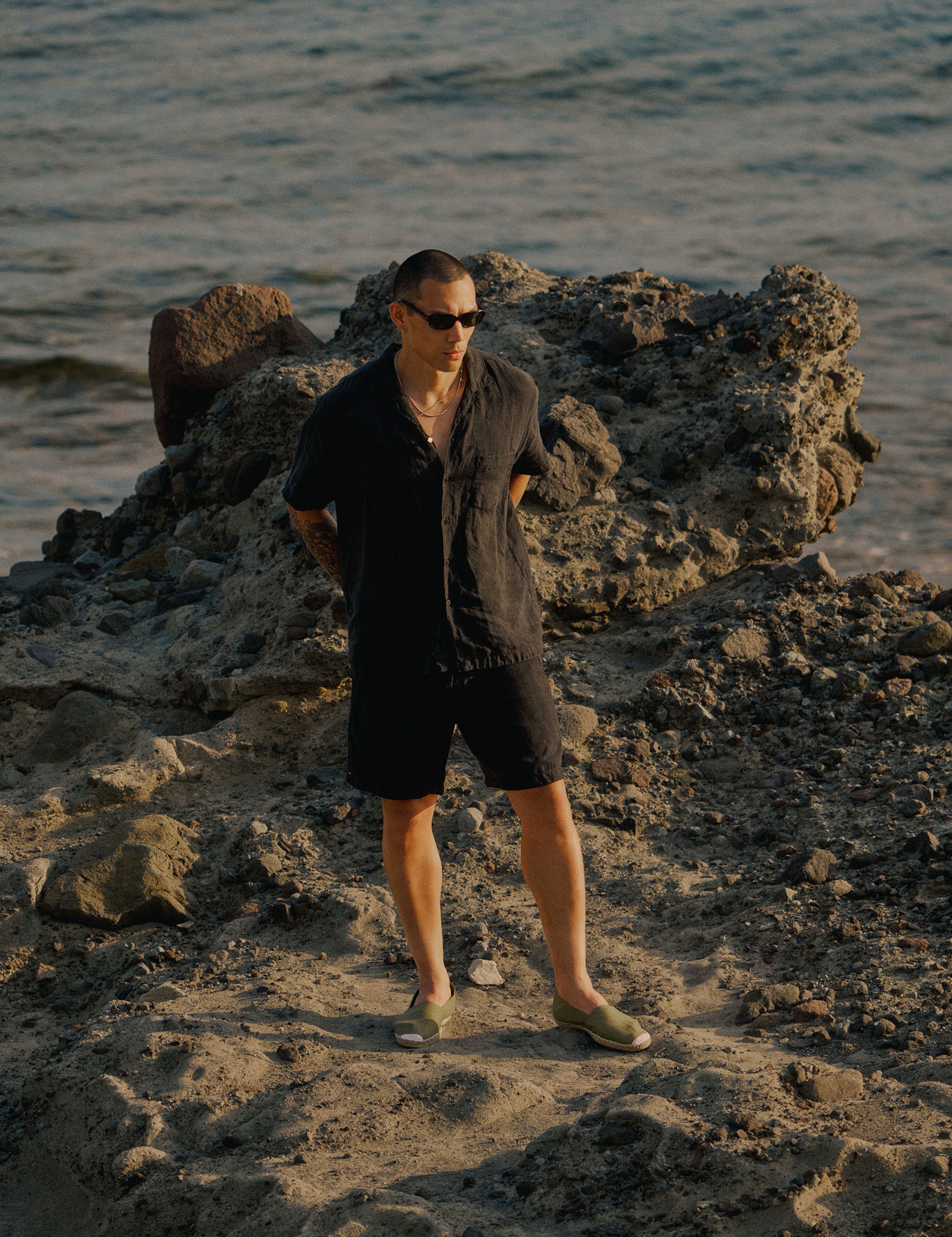 The Holiday Linen SS Shirt in black — worn on a rocky beach by the ocean
