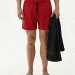 A front view of a person wearing The Shorts in red, holding a black towel. The image highlights the elastic waistband, black drawstrings, and the c'est normal logo on the leg.