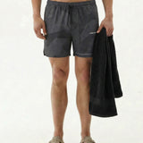 A model wearing The Shorts in Dark Camo, holding a black towel against a white background. The image highlights the shorts' fit, the dark camo pattern, and the white c'est normal logo on the left leg.