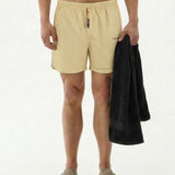 A front view of a shirtless model wearing The Shorts in Lombok Print, standing against a white background holding a black towel. The image highlights the elasticated waistband, black drawstring, and the c'est normal logo on the leg.