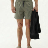 A front view of a model wearing The Shorts in mulled green, standing against a white background. The image highlights the elastic waistband with a dark drawstring, side notches, a small c'est normal logo on the left leg, and the model holding a black towel.