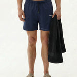 A front view of a model wearing The Shorts in navy against a white background, holding a black towel. The image highlights the elasticated drawstring waistband and the white c'est normal logo on the left leg.