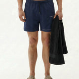 A front view of a model wearing The Shorts in navy against a white background, holding a black towel. The image highlights the elasticated drawstring waistband and the white c'est normal logo on the left leg.