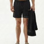 A model standing on a white background wearing the black The Shorts, holding a black towel. The image highlights the c'est normal logo on the left leg.