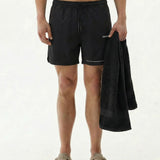 A model standing on a white background wearing the black The Shorts, holding a black towel. The image highlights the c'est normal logo on the left leg.