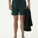A shirtless man standing against a white background, wearing The Shorts in pine green. He holds a black towel in his left hand and is wearing grey slide sandals. The shorts feature the c'est normal logo on the left leg.