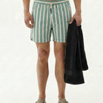 A front view of a model wearing The Shorts in Lombok Stripe on a white background. The image shows the quick-dry, 4-way stretch shorts featuring a drawstring waist, the c'est normal logo on the leg, and the model holding a black towel.