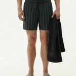 A shirtless model standing against a white background, wearing The Shorts in Phantom Stripe with a dark green and black striped pattern. He holds a black towel in his left hand and wears beige slip-on shoes.